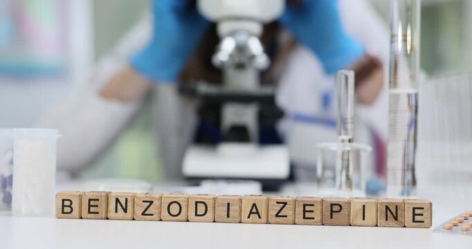 Wooden blocks on desk spell word Benzodiazepine beside glassware. Woman chemist adjusts microscope evaluating tranquilizer drug sample effect