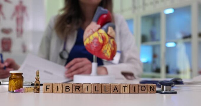 Wooden blocks on desk spell Fibrillation by heart model. Woman cardiologist writes report planning rhythm control therapy at appointment in clinic