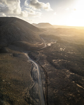 Aerial view of a winding road cutting through the rugged, sun-kissed landscape, with the mountains painted in hues of gold and umber under a radiant sky, Fuerteventura, Canary Islands, Spain.