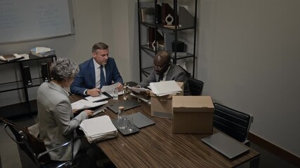 Wide high-angle security camera shot of multiethnic team of defense attorneys sitting around table in meeting room, researching paperwork for precedents while preparing for lawsuit