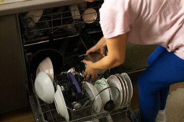 Woman loading dishwasher with plates and utensils, stacking ceramic dishes, placing glasses and silverware, step in evening