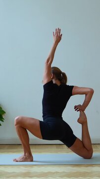 Vertical profile shot of a woman performing Eka Pada Rajakapotasana prep (One-Legged King Pigeon Pose) or a deep low lunge variation. She grips her back foot with her hands, showcasing flexibility