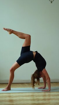 Full vertical shot of a woman in black sportswear performing a deep backbend, likely One-Legged Wheel Pose (Eka Pada Urdhva Dhanurasana). One leg is extended straight up