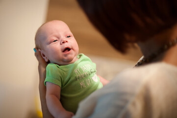 A fair-skinned newborn wearing a green shirt with text is held by an adult with dark hair. The baby gazes upward, its mouth slightly open. The setting is indoors.