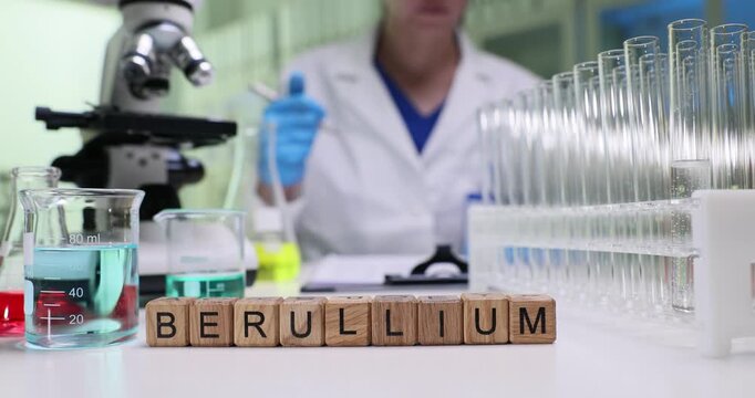 Text Beryllium on wooden cubes among glassware by microscope on scientist desk. Woman chemist fills report about testing results at table in lab
