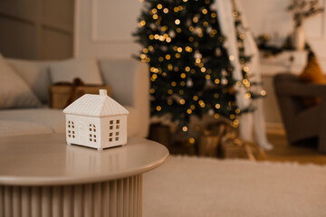 Christmas ceramic house candle on table over xmas tree with glowing lights close up. Winter holiday season. Celebration