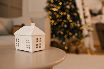 Christmas ceramic house candle on table over xmas tree with glowing lights close up. Winter holiday season. Celebration