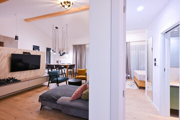 Obraz premium Modern open-plan living and dining area with an integrated kitchen, featuring a comfortable sofa, stylish armchairs, and a bar-height dining table. Wooden floors and ceiling beams enhance this