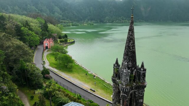 Drone approach to pointed gothic spire of Capela das Vitorias with moss-covered details and Furnas lake backdrop in Azores
