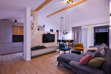 Obraz premium Modern open-plan living and dining area with an integrated kitchen, featuring a comfortable sofa, stylish armchairs, and a bar-height dining table. Wooden floors and ceiling beams enhance this