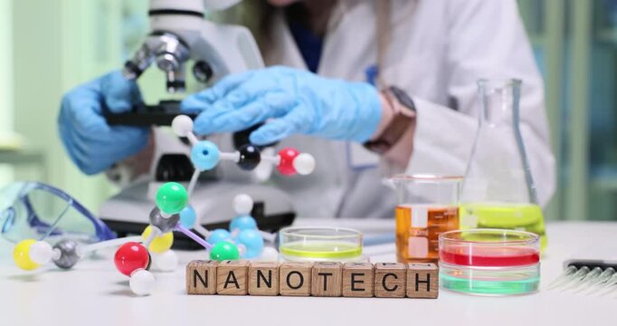 Woman lab technician uses microscope to study delicate structures of sample. Wooden cubes arranged neatly on lab table form word Nanotech at glassware