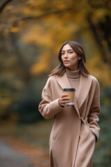 Elegant woman in a beige coat walks through a vibrant autumn park, holding a coffee cup, surrounded by colorful foliage, showcasing a serene outdoor lifestyle