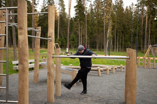 Man doing warm-up exercises outdoors at a fitness park surrounded by forest trees. He stretches his legs on parallel bars, preparing for training and improving flexibility
