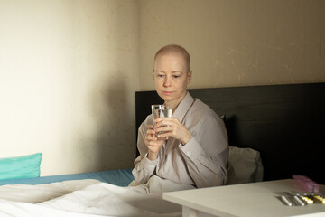 Young woman with shaved head due to cancer therapy resting in bed with glass of water on her lap, comfort and calm atmosphere, chronic illness management, hospital at home style care