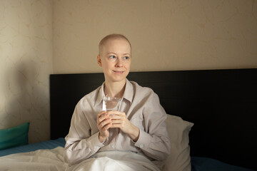 cancer patient resting in bed at home with a glass of water in her hands, bald woman in striped shirt recovering after chemotherapy, concept of oncology treatment, 