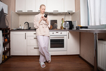 Smiling cancer survivor in soft loungewear standing in modern kitchen and holding smartphone, browsing social media and chatting with friends while appliances and dishes are on the counter, lifestyle