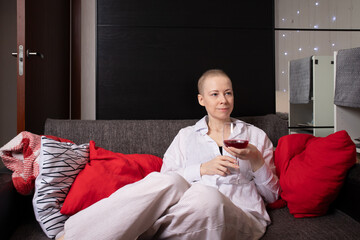 cancer survivor in comfortable striped pajamas reclines on a dark sofa, holding a glass of red drink in her hand and gazing thoughtfully into the distance, 