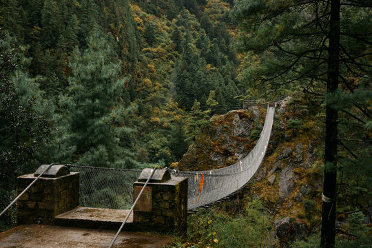 A suspension bridge connects two rocky edges over a deep valley. Trees surround the bridge in autumn colors. The sky is overcast, making the scene look moody
