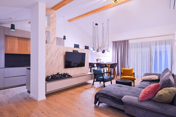 Obraz premium Modern open-plan living and dining area with an integrated kitchen, featuring a comfortable sofa, stylish armchairs, and a bar-height dining table. Wooden floors and ceiling beams enhance this