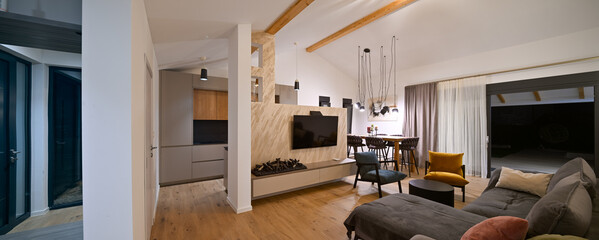 Obraz premium Modern open-plan living and dining area with an integrated kitchen, featuring a comfortable sofa, stylish armchairs, and a bar-height dining table. Wooden floors and ceiling beams enhance this