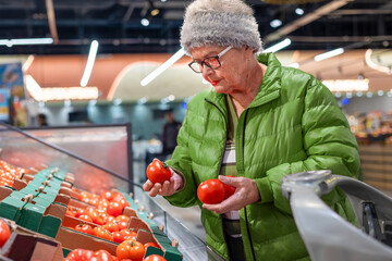 Senior woman comparing and selecting fresh tomatoes in produce section of modern supermarket. healthy eating, fresh food selection, independent senior lifestyle, mindful grocery shopping and consumer 