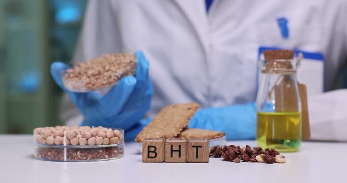 Wooden blocks on lab desk spell code BHT near nuts and oil in bottle. Scientist in gloves holds cereal cracker sample studying antioxidant effect
