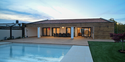Luxurious modern villa at twilight, featuring a serene swimming pool reflecting warm interior lights. An inviting wooden deck hosts an outdoor dining area, complemented by a stylish hanging chair