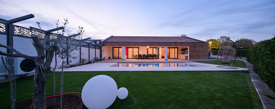 Luxurious modern villa at twilight, featuring a serene swimming pool reflecting warm interior lights. An inviting wooden deck hosts an outdoor dining area, complemented by a stylish hanging chair