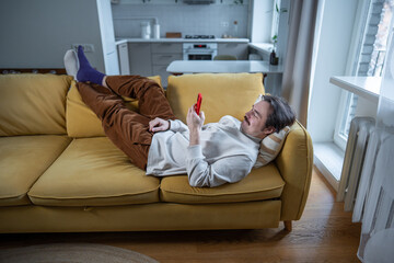 Smiling relaxed man with phone lying on sofa surfing online web, procrastination at home. Lazy male with social media addiction on smartphone scrolling internet, enjoy virtual life looking at screen. 