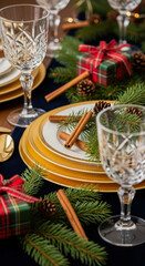 Festive Christmas table setting with golden plates, crystal glasses, cinnamon sticks and plaid ribbon gifts on a navy tablecloth.