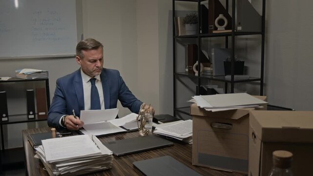 Medium full shot of middle-aged Caucasian male barrister in blue suit and tie sitting at desk alone in office, studying piles of documents while preparing arguments and strategy for court trial
