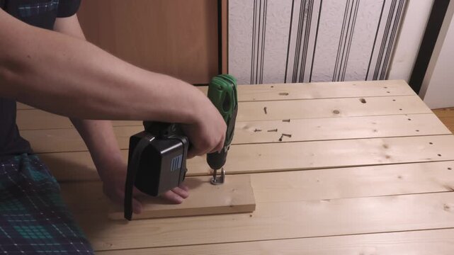 Unrecognizable adult uses a cordless drill to mount a metal bracket on a wood plank, screws scattered on the table. DIY home improvement scene