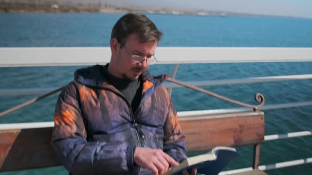white man reading book on bench by sea, puffer jacket, glasses, turning pages, calm waves, bright sky, waterfront promenade, pensive expression, retired teacher revising notes, morning light