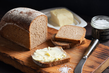Square loaf of artisan rye bread cut into slices served with butter and sea salt on wooden cutting board