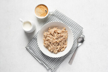 Healthy vegan breakfast meal: cooked oatmeal and spelt porridge in white bowl served with cup of coffee and almond milk in jug