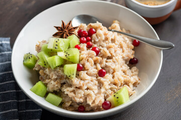 Oatmeal porridge with kiwi and cranberries in white ceramic bowl on wooden table, closeup view. Healthy breakfast meal rich in dietary fiber