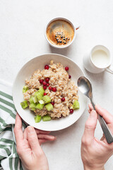 Healthy breakfast concept: woman hands holding bowl of oatmeal porridge with kiwi, cranberries and cup of coffee on side. Top view. Diet meal