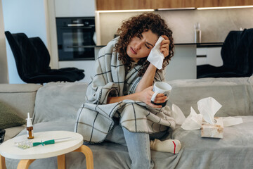 Fototapeta premium White cup is in hand. Sick woman feeling unwell at home, checking her temperature and using medicine while resting on a sofa