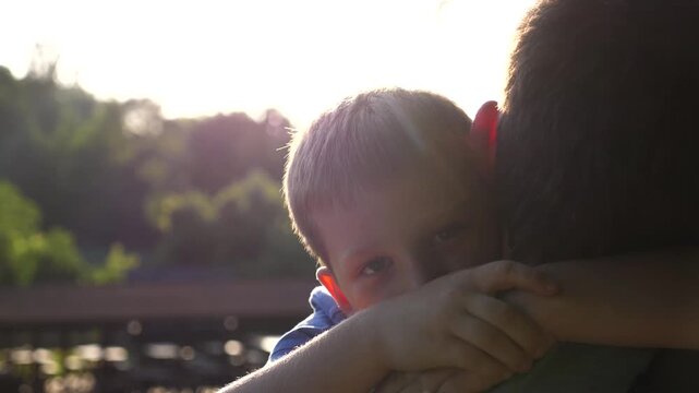 Young dad holding in hands his small son at countryside. Little boy looking into camera and hugging his father against sunlight at background. Concept of family happiness. Close up Slow motion