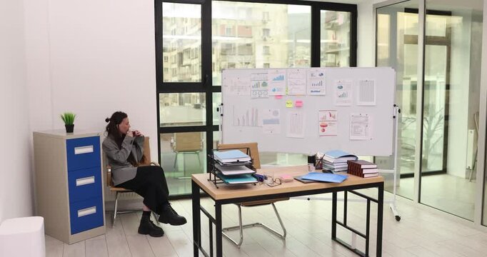 Lady office worker sitting near white board drinks hot tea. Woman relaxes in bright office before presentation while waiting for business meeting