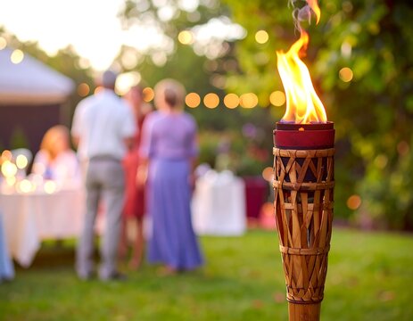 Lit tiki torch in a backyard with blurred partygoers and soft evening light in a summery outdoor scene