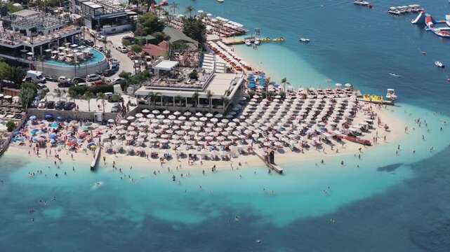Aerial view of Ksamil's vibrant beach, dotted with umbrellas, boats on turquoise waters, and coastline buildings, creating a scenic landscape, Ksamil, Vlore County, Albania.