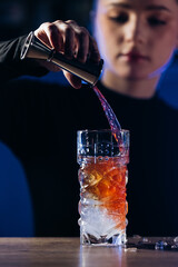 Female bartender skillfully pouring a vibrant cocktail into a glass filled with ice, showcasing mixology techniques in a modern bar environment with ambient lighting