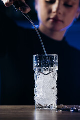 Female bartender pouring liquid into a glass filled with ice, showcasing cocktail preparation skills in a dimly lit bar environment with vibrant colors