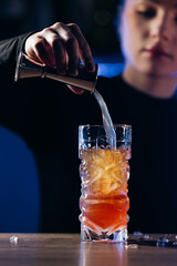 Bartender pouring a colorful cocktail into a glass filled with ice, showcasing mixology skills and vibrant drink presentation in a stylish bar environment