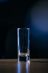Clear glass shot glass stands upright on a dark surface, illuminated by soft blue light, showcasing its sleek design and reflective qualities in a minimalist setting