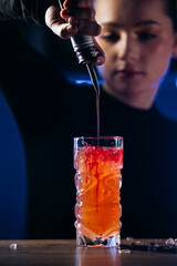 Female bartender skillfully pouring syrup into a vibrant cocktail glass, showcasing colorful layers and textures, with a blurred background creating an inviting atmosphere