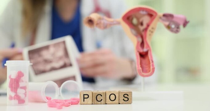 Wooden blocks on gynecology desk spell word PCOS while female doctor studies ultrasound scan. Uterus model with pills symbolize hormone disorder