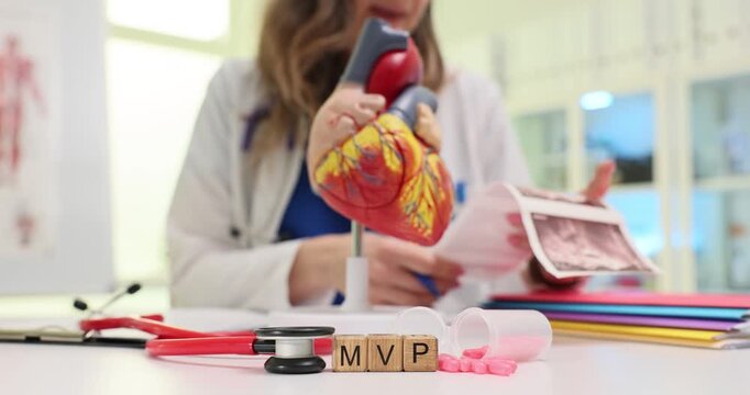 Wooden blocks on cardiological table spell MVP while female cardiologist holds scan images. Doctor reviews echocardiogram printout about mitral valve