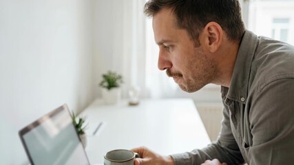 Focused young man working at home office desk while drinking coffee and concentrating on laptop screen in bright minimal workspace, 4k video